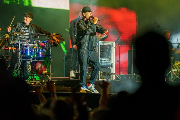 Musiker mit Instrumenten auf der Bühne, Publikum hebt die Hände.